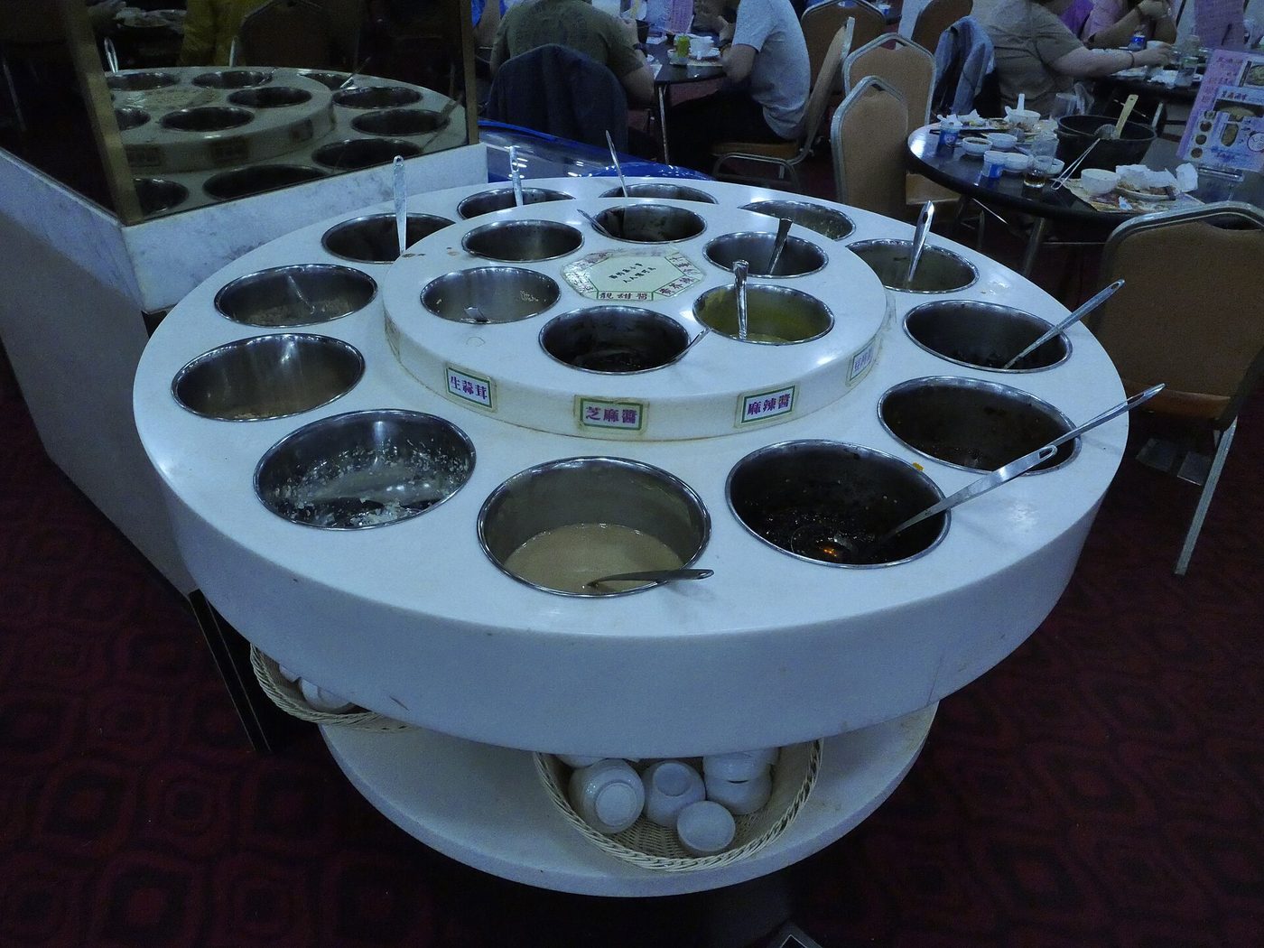 A hotpot condiment station with separate bowls, serving spoons and a sesame-paste label